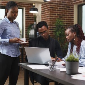 Financial company employees reviewing marketing strategy documentation and development plan. African team leader assistant holding a cup of coffee while standing near office workspace desk