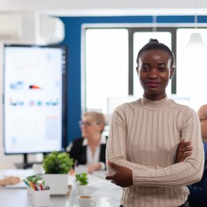 Successful smiling african business woman holding arms crossed looking atcamera in conference room. Manager working in professional start up financial business, modern company workplace ready for meeting.