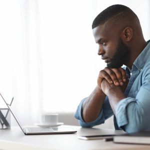 Thoughtful Black Writer Working With Laptop At Home Office. African American Freelancer Man Sitting At Desk And Looking At Computer Screen With Pensive Face Expression, Side View With Copy Space